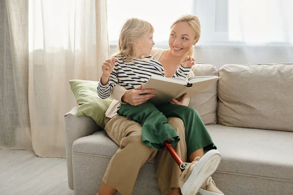 Glückliches Kind mit Beinprothese sitzt auf dem Schoß einer blonden Mutter und liest Buch im Wohnzimmer — Stockfoto