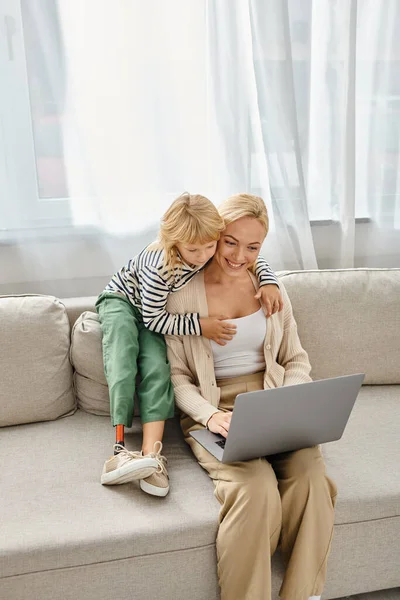 Happy child with prosthetic leg hugging blonde mother working on laptop in modern living room — Stock Photo