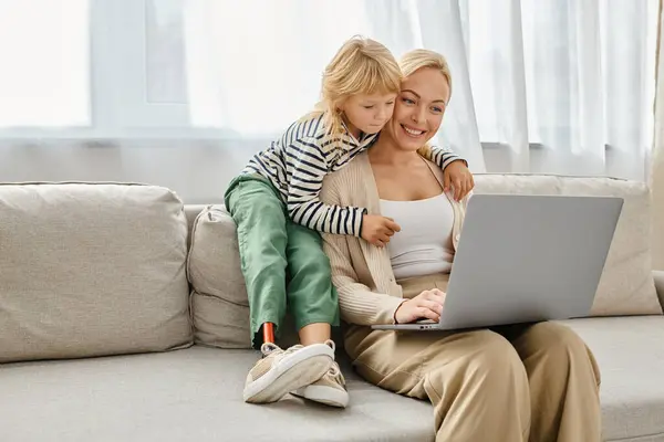 Happy girl with prosthetic leg hugging blonde mother working on laptop in modern living room — Stock Photo
