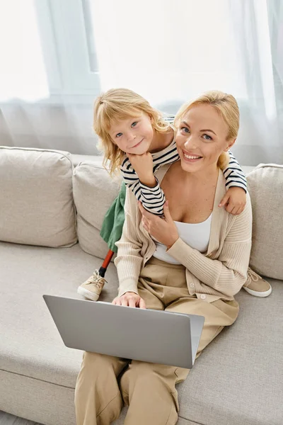 Маленькая девочка с протезной ногой обнимает счастливую мать работает на ноутбуке в современной гостиной — стоковое фото