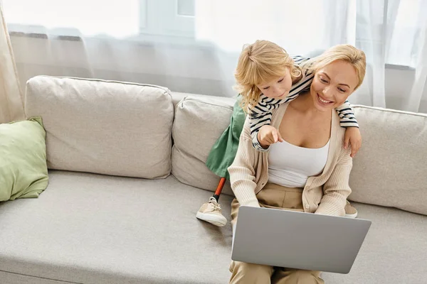 Enfant heureux avec prothèse jambe étreignant mère blonde travaillant sur ordinateur portable dans le salon moderne — Photo de stock