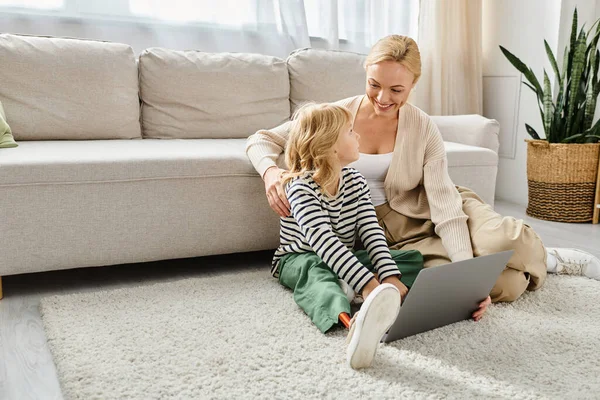 Niño feliz con la pierna protésica usando el ordenador portátil y sentado en la alfombra con la madre en la sala de estar - foto de stock