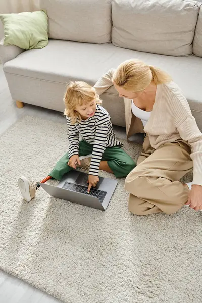 Niño feliz con la pierna protésica apuntando al ordenador portátil y sentado en la alfombra con la madre en la sala de estar - foto de stock