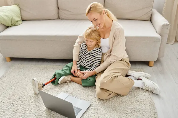 Happy child with prosthetic leg watching movie on laptop while sitting on carpet with mother — Stock Photo