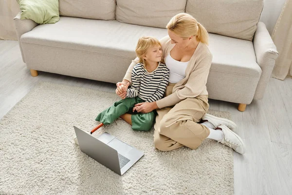 Chica feliz con la pierna prótesis viendo la película en el ordenador portátil y sentado en la alfombra junto con mamá - foto de stock