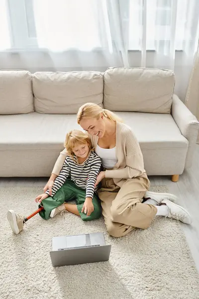 Niño feliz con la pierna prótesis viendo la película en el ordenador portátil y sentado en la alfombra junto con la madre - foto de stock