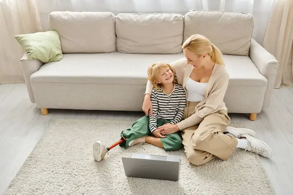 Fille heureuse avec prothèse jambe regarder film sur ordinateur portable et assis sur le tapis avec la mère — Photo de stock