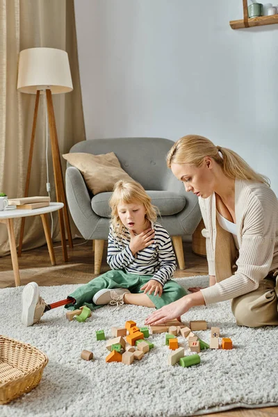 Chica confundida con la pierna protésica sentado en la alfombra y mirando los juguetes de madera cerca de la madre - foto de stock
