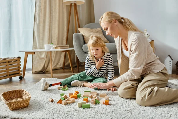 Disabled girl with prosthetic leg sitting on carpet and looking at wooden toys near mother — Stock Photo