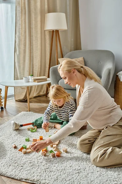 Bambina disabile con gamba protesica seduta su tappeto e che gioca con giocattoli in legno vicino alla madre — Foto stock
