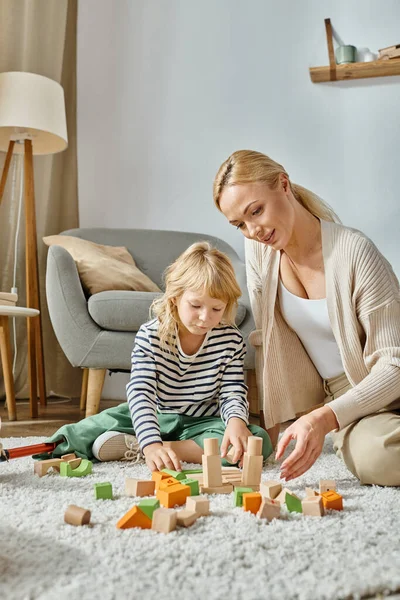 Маленькая девочка с протезной ногой, сидящая на ковре и играющая с деревянными игрушками рядом с матерью блондинкой — стоковое фото