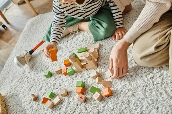 Chica recortada con la pierna protésica sentado en la alfombra y jugando con juguetes de madera cerca de la madre - foto de stock