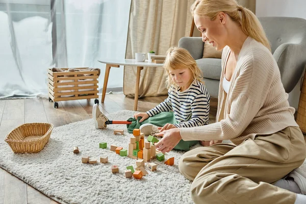 Little girl with prosthetic leg sitting on carpet and playing with wooden toys near joyful mother — Stock Photo