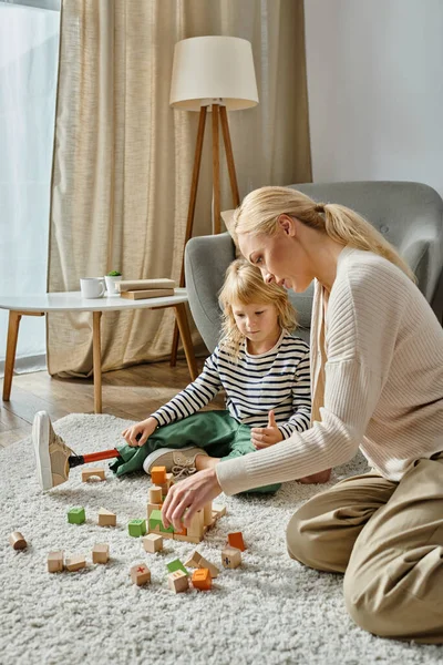 Disabled girl with prosthetic leg sitting on carpet and playing with wooden toys near blonde mother — Stock Photo