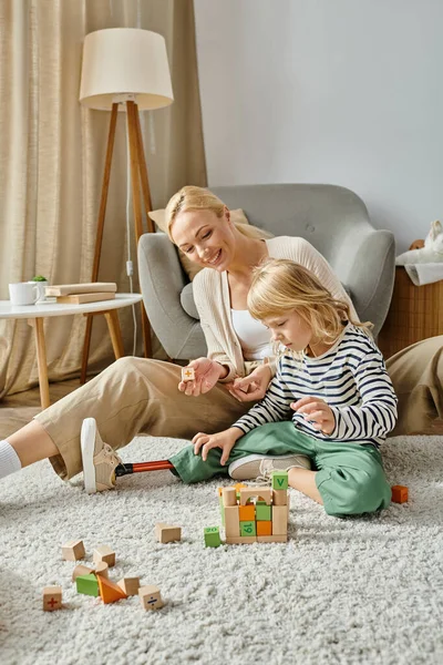 Niña con la pierna protésica sentada en la alfombra y jugando con juguetes de madera cerca de la madre alegre - foto de stock