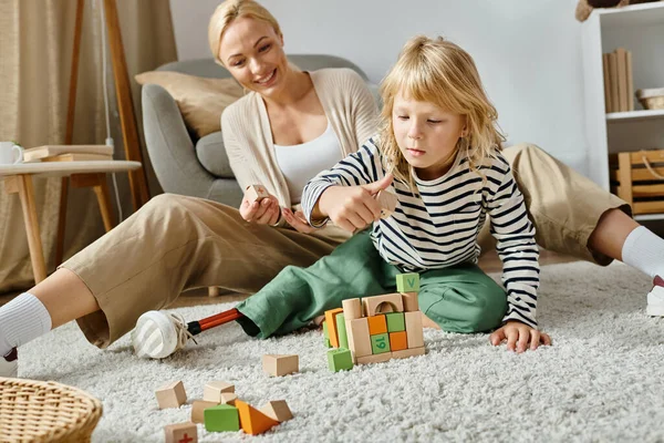 Niña con la pierna protésica sentada en la alfombra y jugando con bloques de madera cerca de la madre feliz - foto de stock