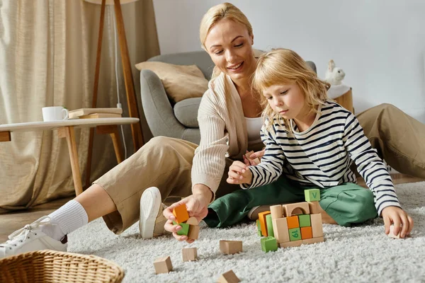 Cure girl with prosthetic leg sitting on carpet and playing with wooden blocks near happy mother — Stock Photo