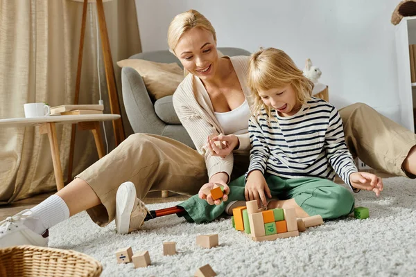 Preschool girl with prosthetic leg sitting on carpet and playing with wooden blocks near mother — Stock Photo
