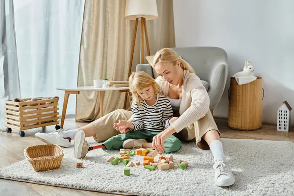 Chica rubia con la pierna protésica sentado en la alfombra y jugando con bloques de madera cerca de la madre en casa - foto de stock