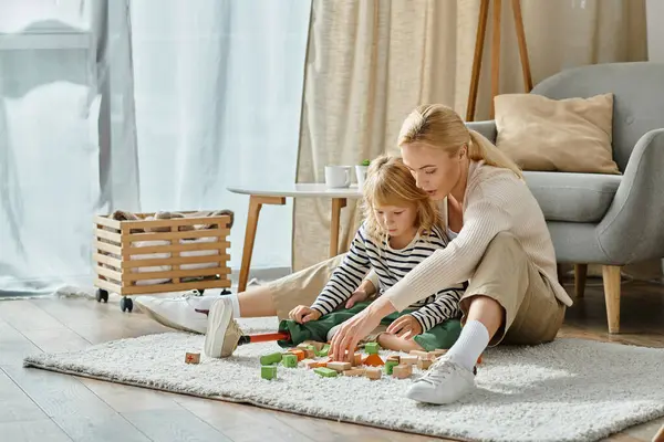 Chica rubia con la pierna protésica sentado en la alfombra y jugando bloques de madera juego cerca de la madre cariñosa - foto de stock