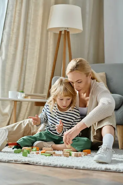 Blonde girl with prosthetic leg sitting on carpet and playing with wooden blocks near caring mom — Stock Photo