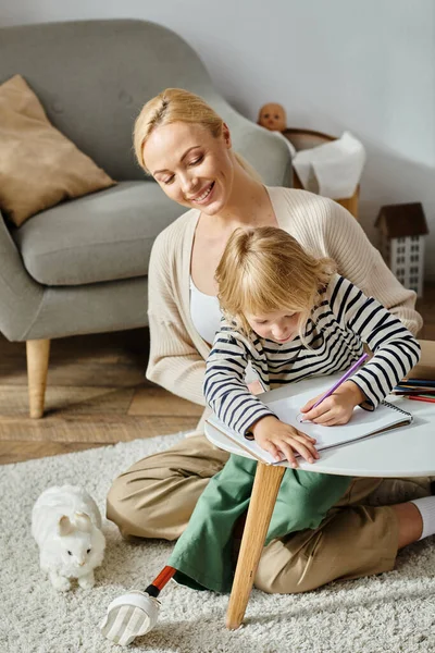 Blondes Mädchen mit Beinprothese auf Papier mit Buntstiften in der Nähe der glücklichen Mutter zu Hause — Stockfoto