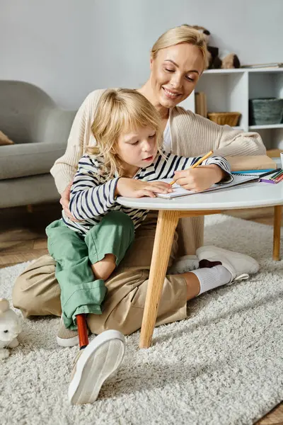 Happy blonde woman looking at her daughter with prosthetic leg drawing on paper with colorful pencil — Stock Photo