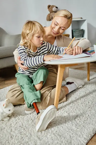 Madre feliz mirando a su hija con el dibujo de la pierna protésica en papel con lápiz de colores - foto de stock