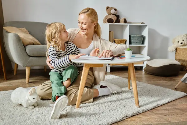 Happy blonde woman looking at her daughter with prosthetic leg drawing on paper with colorful pencil — Stock Photo
