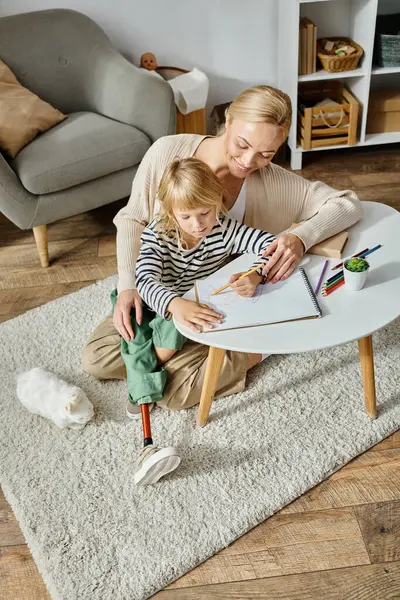 Счастливая блондинка смотрит на свою дочь с протезной ногой, рисующей на бумаге цветным карандашом — стоковое фото