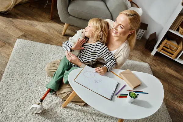 Счастливая женщина обнимает дочь с протезной ногой рядом с бумагой и красочным карандашом на столе, над головой — стоковое фото