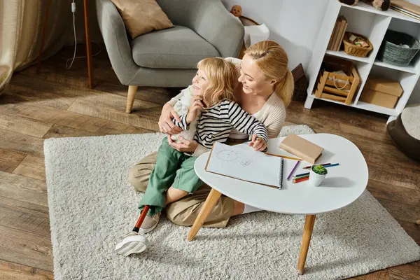 Woman hugging happy daughter with prosthetic leg near paper and colorful pencil on table, overhead — Stock Photo