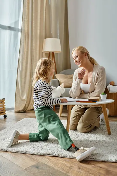 Madre felice guardando figlia con gamba protesica seduta vicino al tavolo con disegno e peluche — Foto stock