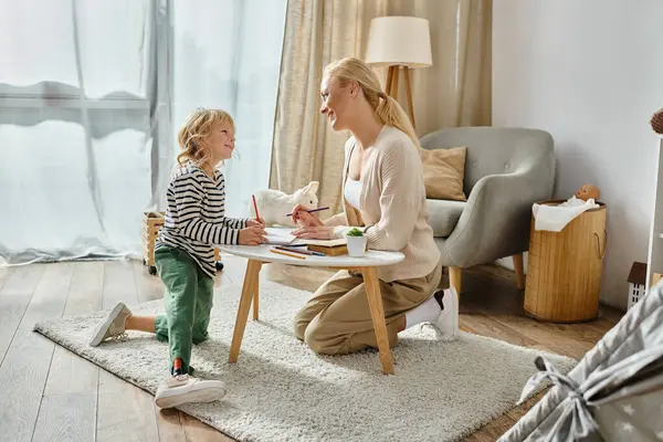 Счастливая мать смотрит на своего ребенка с протезной ногой, рисующей на бумаге цветным карандашом — стоковое фото
