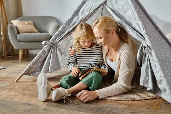 Mignonne fille avec prothétique jambe livre de lecture et assis avec mère heureuse dans la tente de jeu à la maison — Photo de stock