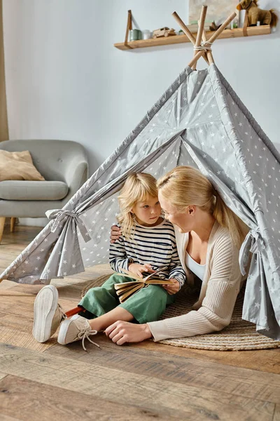 Mignonne fille avec prothétique jambe livre de lecture et assis avec mère heureuse dans la tente de jeu à la maison — Photo de stock