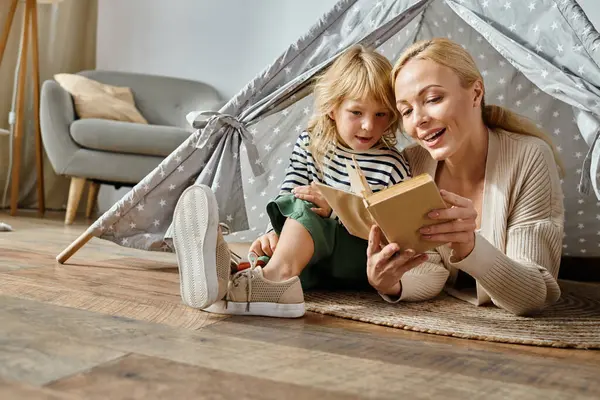 Carino ragazza con protesi gamba e bionda madre lettura libro e seduto in tenda da gioco a casa — Foto stock