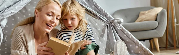 Bonito preteen menina e loira mãe leitura livro e sentado juntos em jogar tenda em casa, banner — Fotografia de Stock