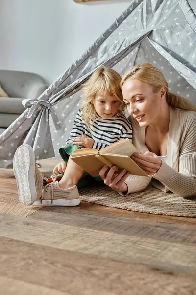 Nettes Mädchen mit Beinprothese und blonder Mutter, die Buch liest und zu Hause im Spielzelt sitzt — Stockfoto