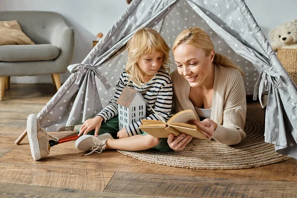 Bonito menina com prótese perna e loira mãe leitura livro e sentado em jogar tenda em casa — Fotografia de Stock