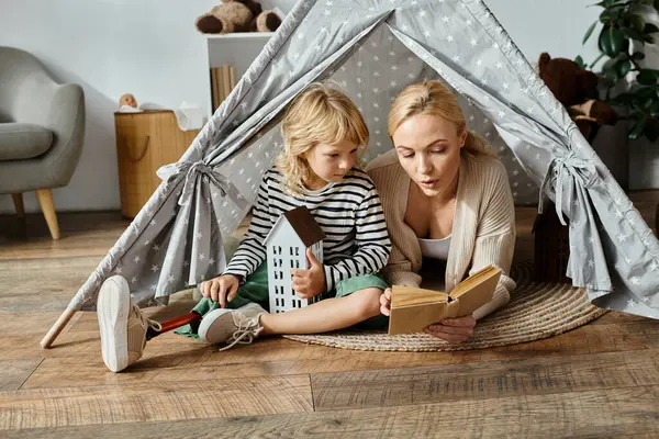 Fille mignonne avec prothèse jambe et blonde mère lecture livre tout en étant assis dans la tente de jeu à la maison — Photo de stock