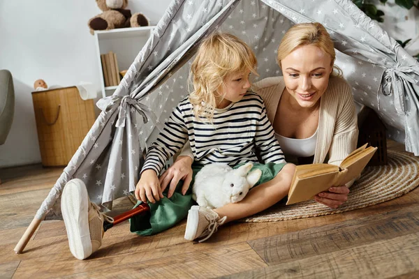Kleines Mädchen mit Beinprothese hält Kuscheltier in der Nähe von Mutter beim Lesen von Buch im Spielzelt zu Hause — Stockfoto