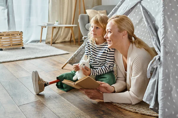 Dreamy girl with prosthetic leg and mother looking away and reading book while sitting in play tent — Stock Photo