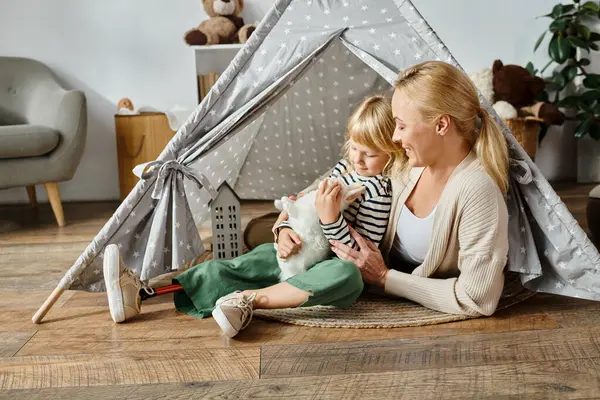 Little girl with prosthetic leg hugging soft toy rabbit near happy mother while sitting in play tent — Stock Photo