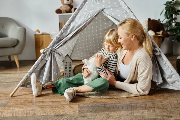 Kleines Mädchen mit Beinprothese umarmt Stofftier-Kaninchen neben glücklicher Mutter, während es im Spielzelt sitzt — Stockfoto