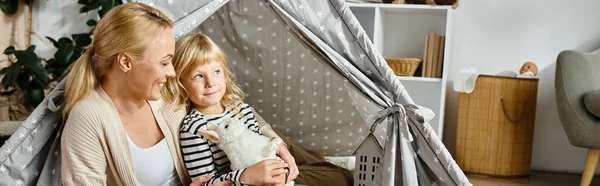 Bonito menina segurando macio brinquedo coelho perto feliz mãe enquanto sentado juntos no jogar tenda, banner — Fotografia de Stock