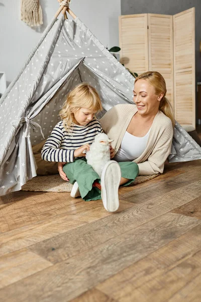Fille blonde avec prothèse jambe jouer avec doux jouet lapin près de mère tout en étant assis dans la tente de jeu — Photo de stock