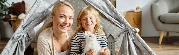 Bannière de portrait de famille, fille heureuse tenant un jouet doux près de la mère dans la tente de jeu et regardant la caméra — Photo de stock