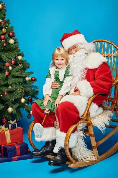Happy girl with prosthetic leg sitting on laps of Santa Claus next to Christmas tree on blue — Stock Photo