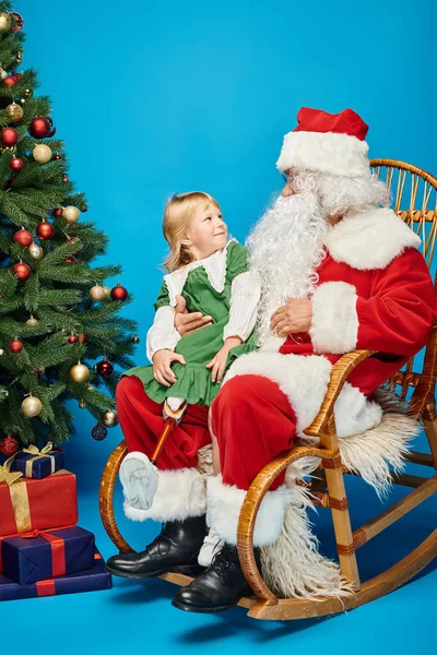 Chica feliz con la pierna protésica sentado en las vueltas de Santa Claus al lado del árbol de Navidad en azul - foto de stock